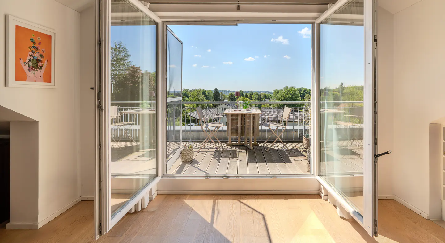 Open glass doors lead to a balcony with a table and chairs, overlooking a green landscape under a blue sky. Interior has light wood floors and white walls.