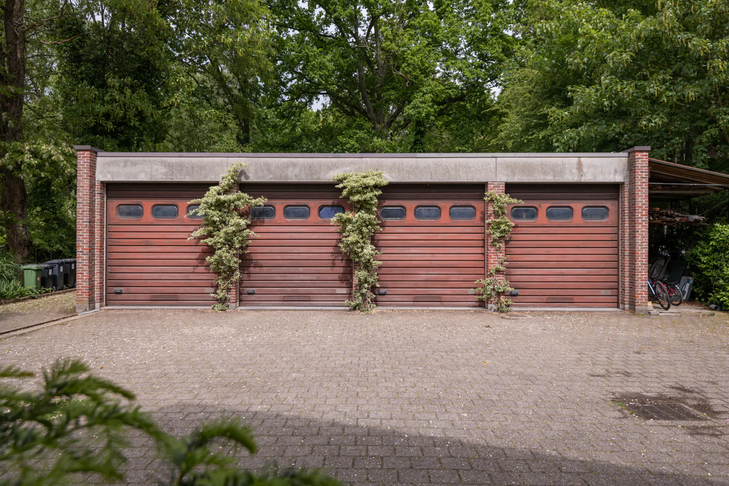 A detached brick garage with four brown sectional doors, accented by climbing vines, set against a backdrop of lush trees.