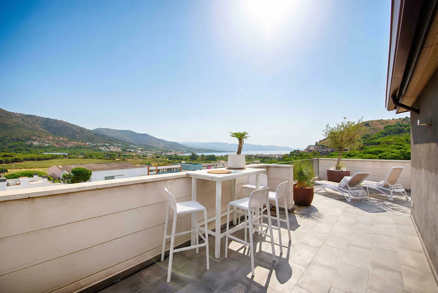 Rooftop patio with white table, chairs, and lounge chairs overlooking a green landscape and blue sky.