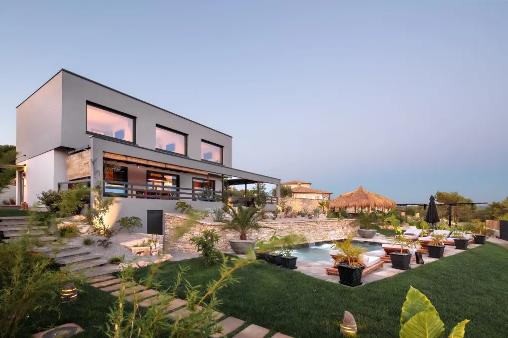 Modern two-story home with a pool and tiki hut. The house is gray and white with large windows. The pool is surrounded by lounge chairs and plants.