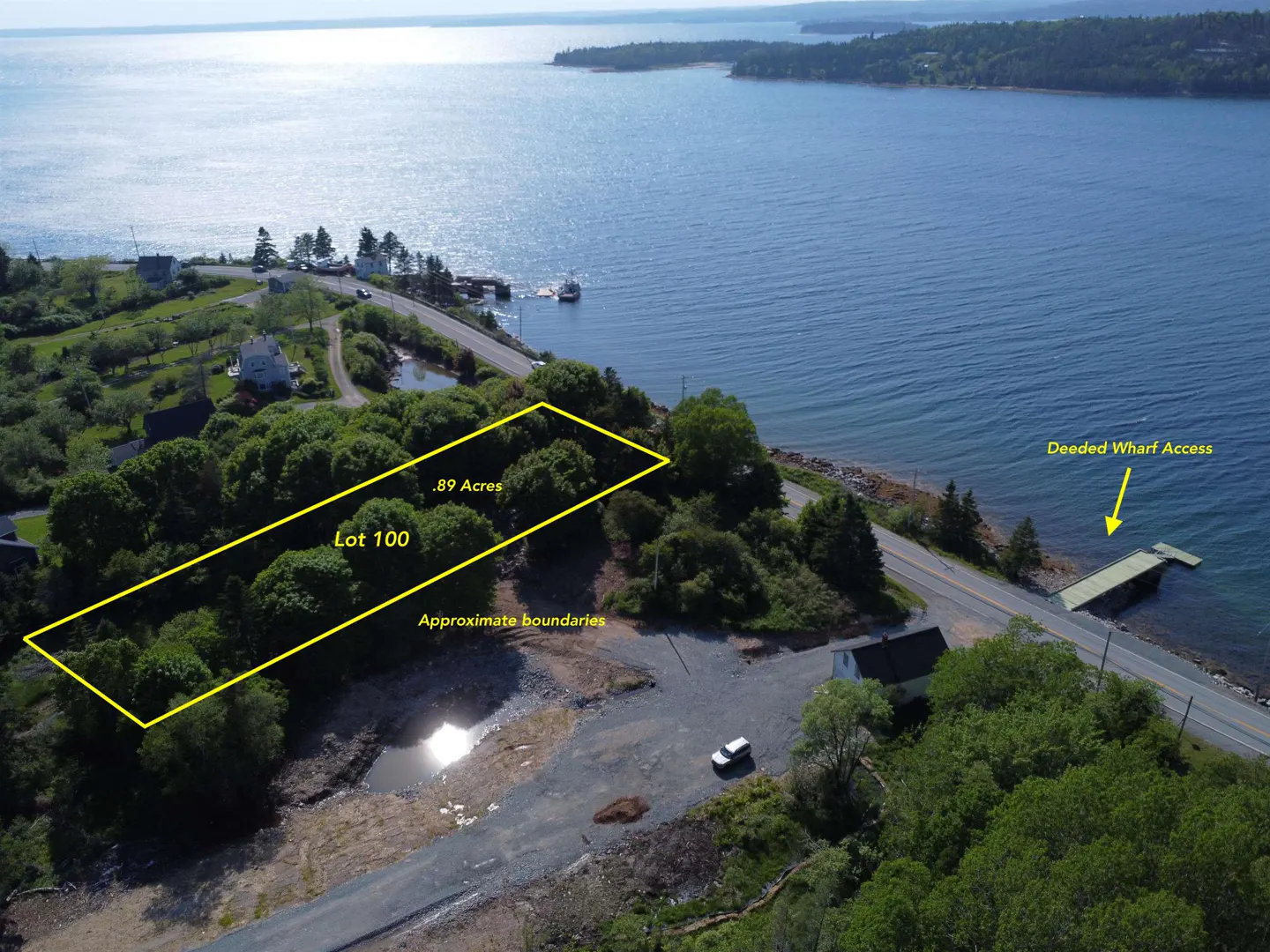 Aerial view of Lot 100, .89 acres, outlined in yellow, near a road, wharf, and blue ocean. Trees surround the lot.