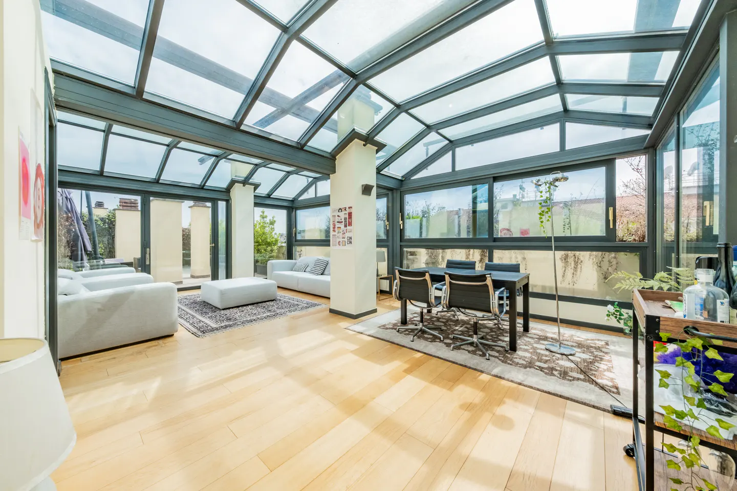 Bright sunroom with glass ceiling, light wood floors, white sofas, and a black dining table with four chairs.