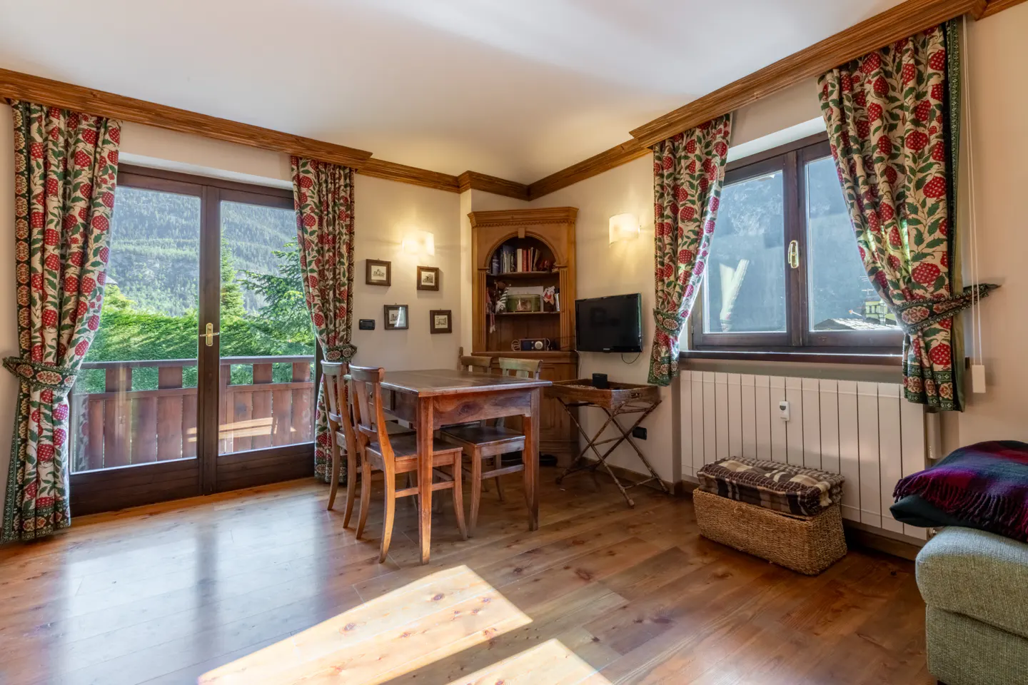 Cozy living room with wood floors, a dining table, and floral curtains framing a balcony view of lush greenery.