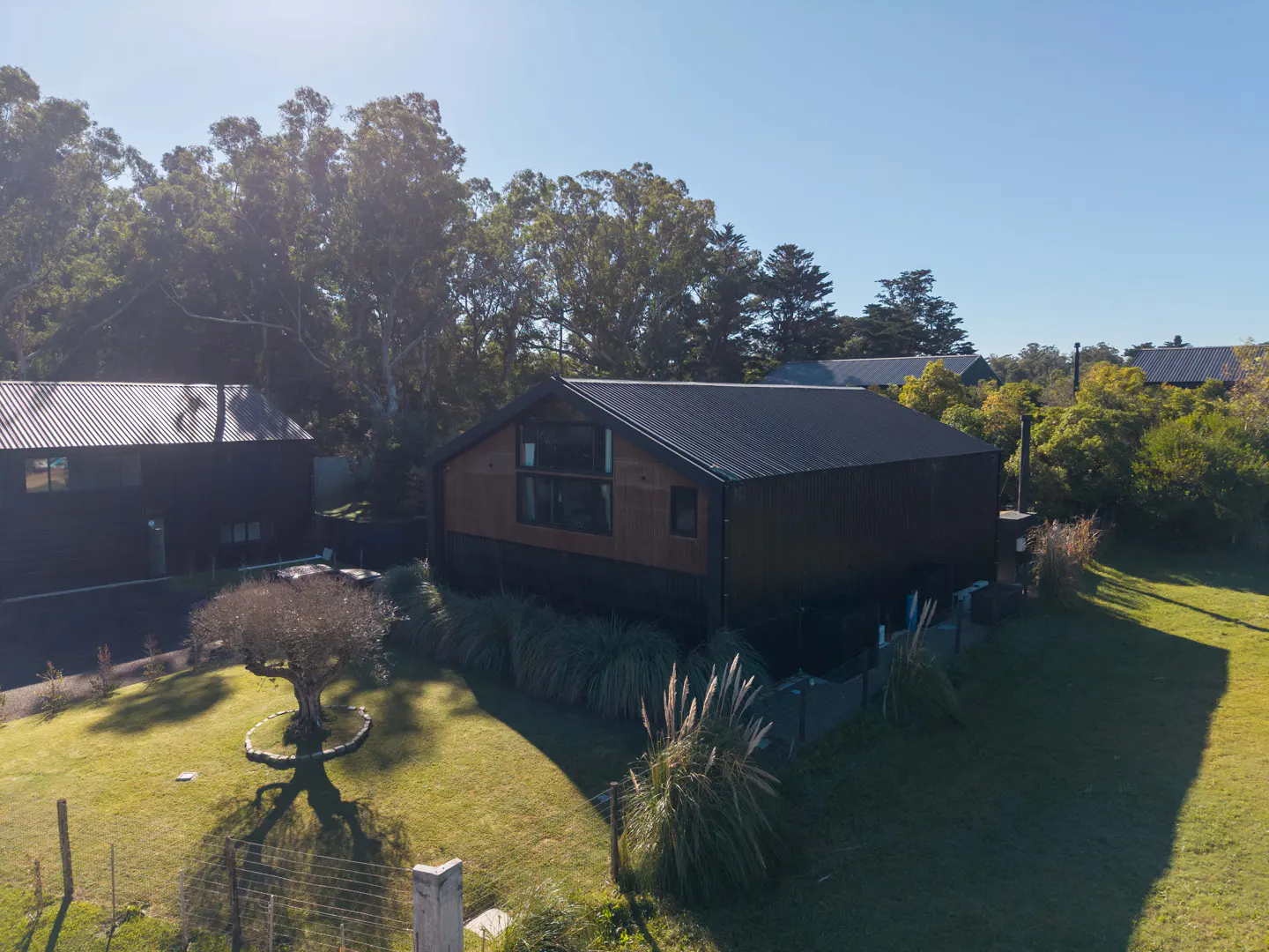 Aerial view of a modern, dark brown house with a black metal roof, surrounded by green grass and trees under a clear blue sky.