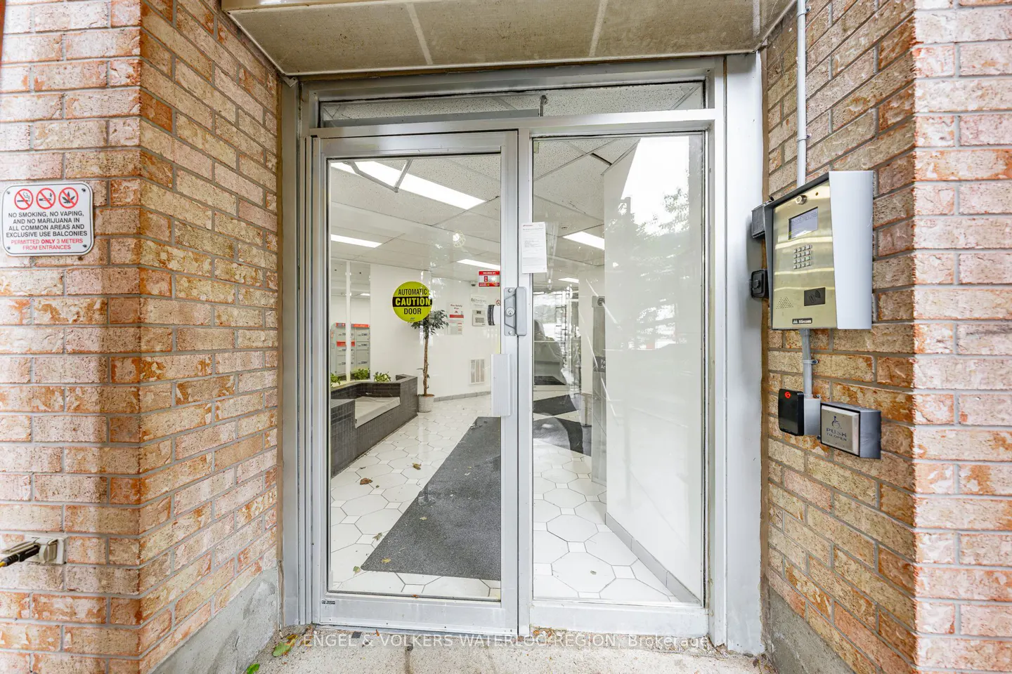 Exterior view of a building entrance with brick walls and a glass door. An intercom system is mounted on the right wall.