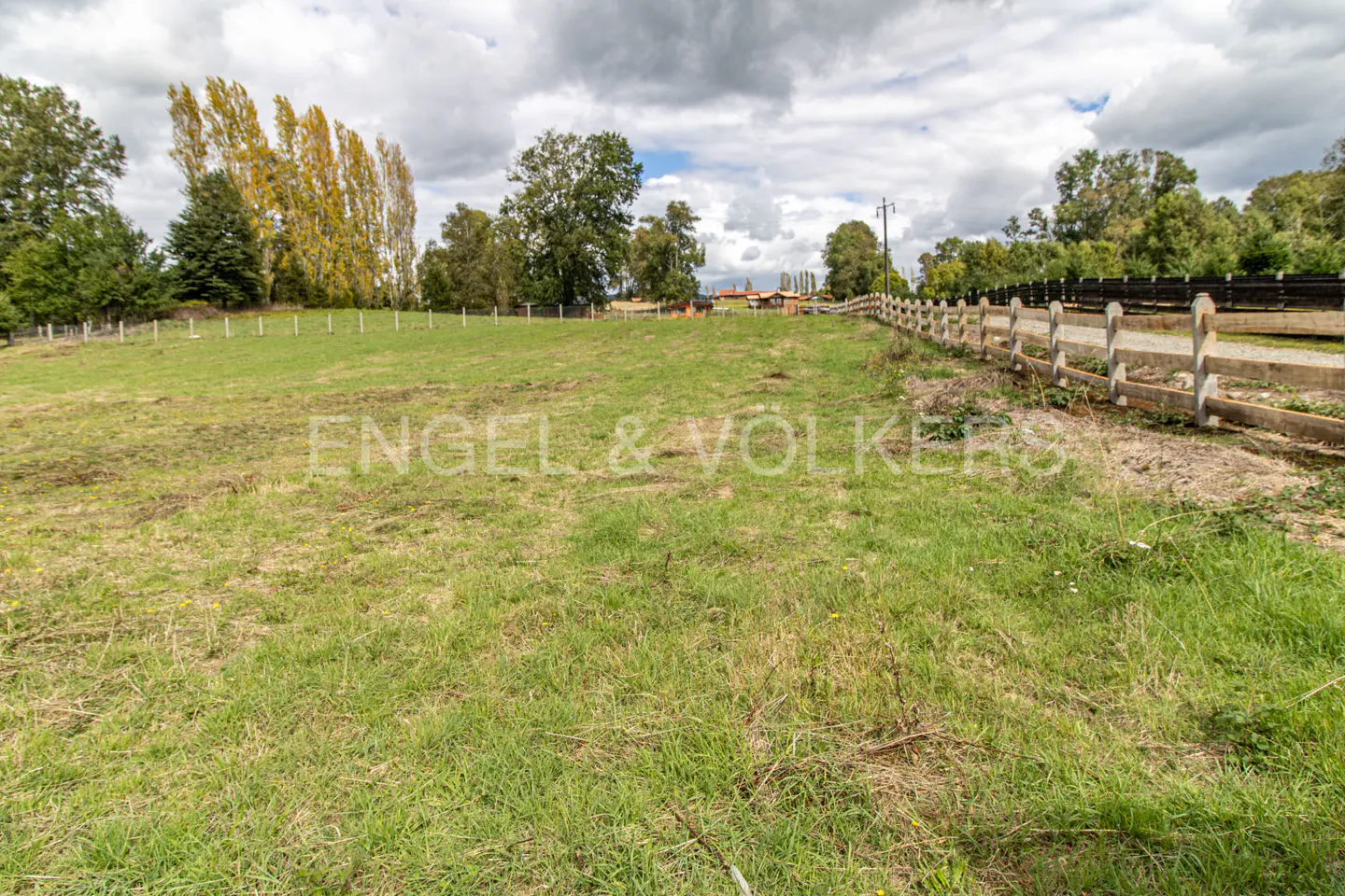 A grassy field with a wooden fence on the right side, trees in the background, and a cloudy sky above.