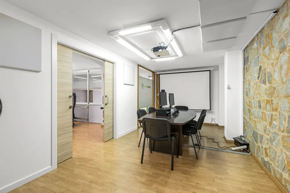 Conference room with a dark table, black chairs, projector, and screen. One wall is stone, and a sliding door leads to another office.