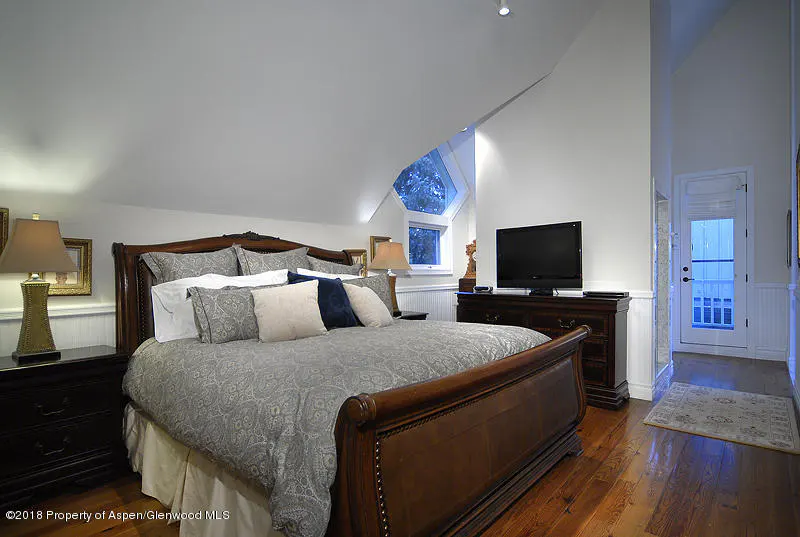 Bedroom with a wooden bed frame, gray patterned bedding, and several pillows. A TV sits on a dark wood dresser. A window is visible in the background.