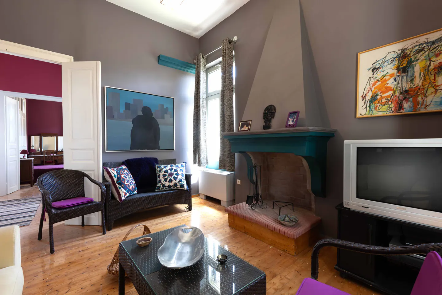 Living room with gray walls, wood floors, and colorful accents. A teal fireplace, modern art, and wicker furniture add character.