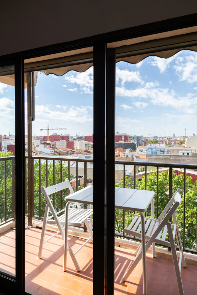Balcony view through sliding glass doors. White table and chairs sit on a terracotta-tiled floor, overlooking a cityscape under a blue, cloudy sky.