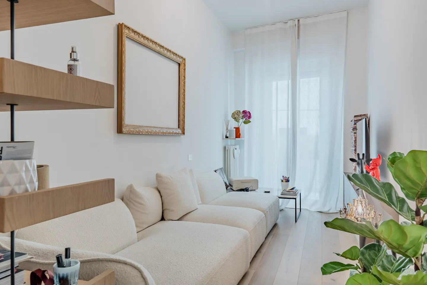 Modern living room with a cream sectional sofa, gold-framed art, sheer curtains, and a large green plant in the foreground.
