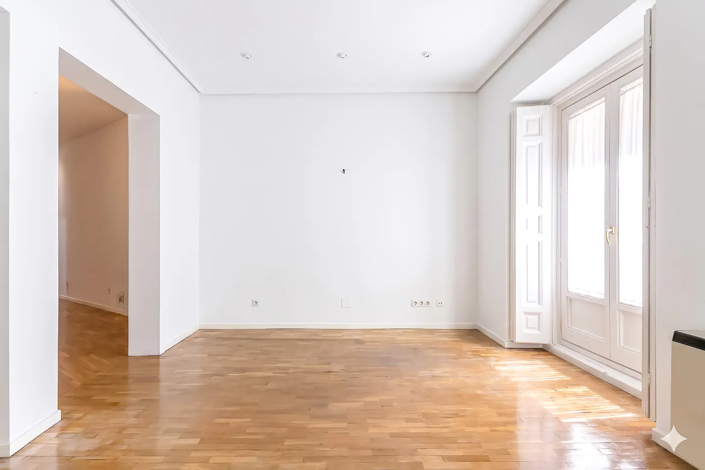 Bright, empty room with white walls, hardwood floors, and a large window with white shutters. An open doorway leads to another room.