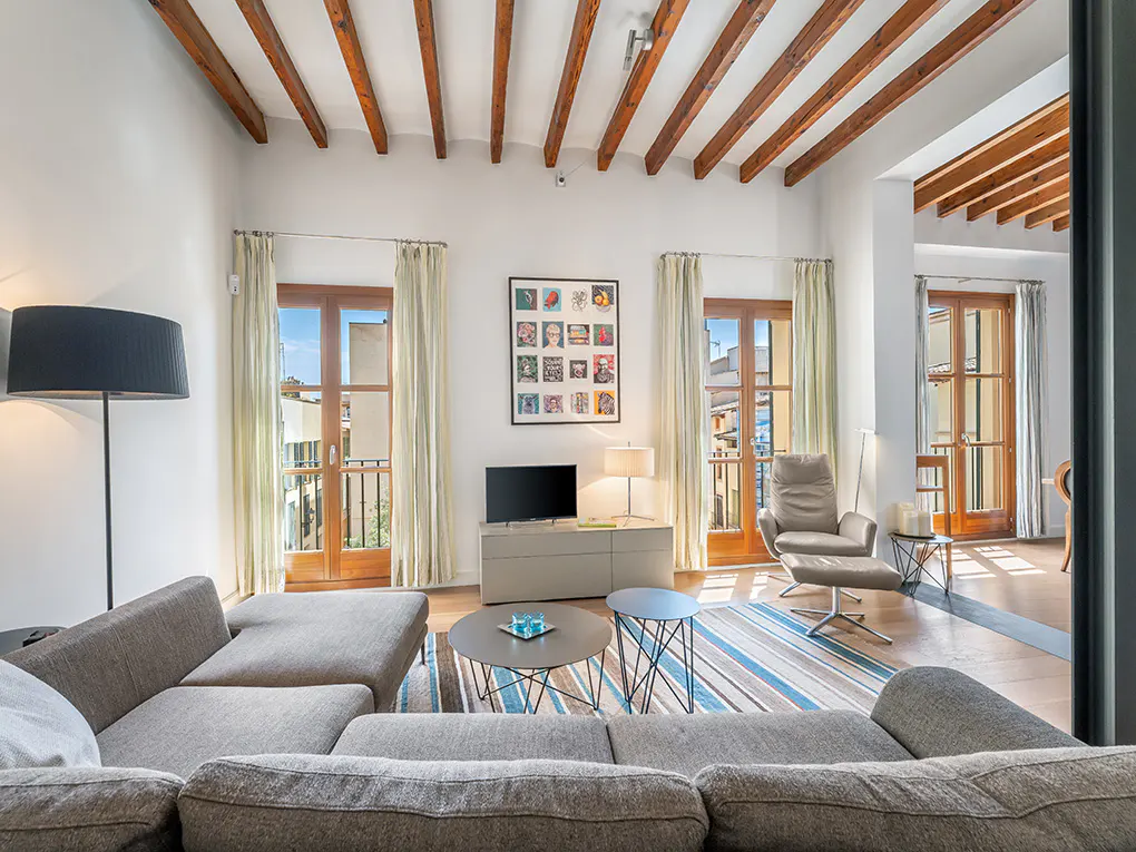 Living room with exposed wood beams, gray sectional sofa, striped rug, and two sets of French doors leading to balconies.
