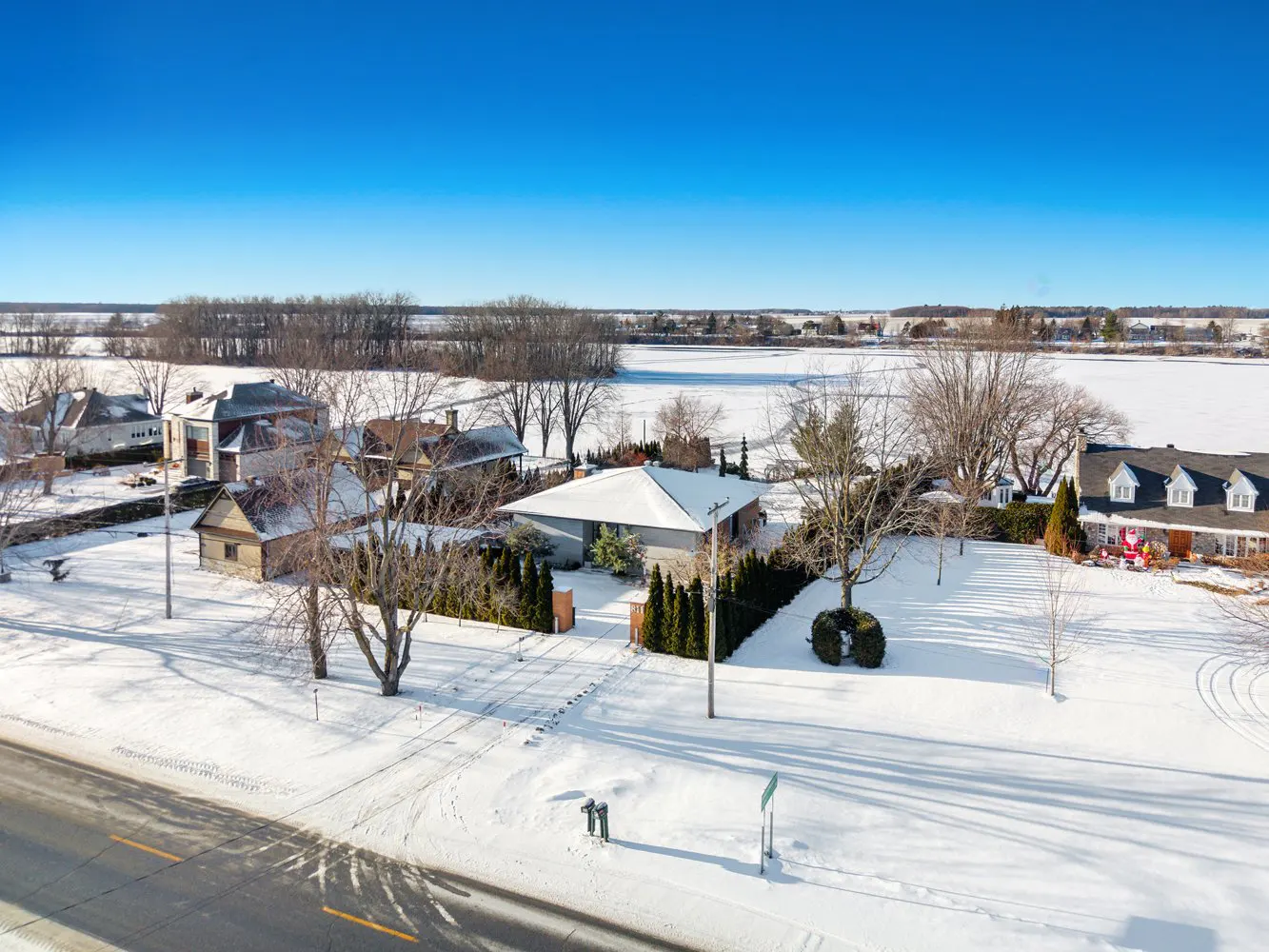 Aerial view of houses covered in snow under a clear blue sky. Trees are bare, and a road runs in front of the properties.