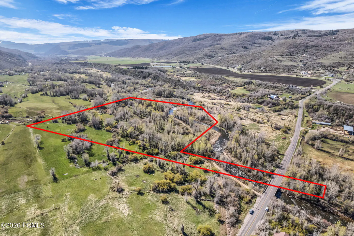 Aerial view of a large, rectangular plot of land outlined in red, with a river running through it, surrounded by green fields and mountains.