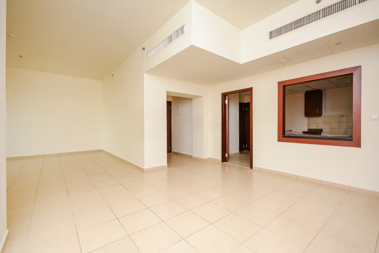 An empty room with beige tile flooring and white walls. A doorway and a window to the kitchen are visible.