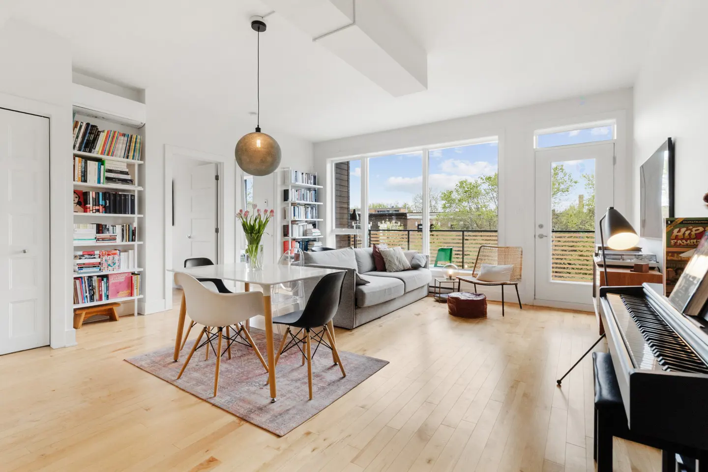 Bright, open living space with hardwood floors, a dining table, and a gray sofa near a large window. Bookshelves line the walls.