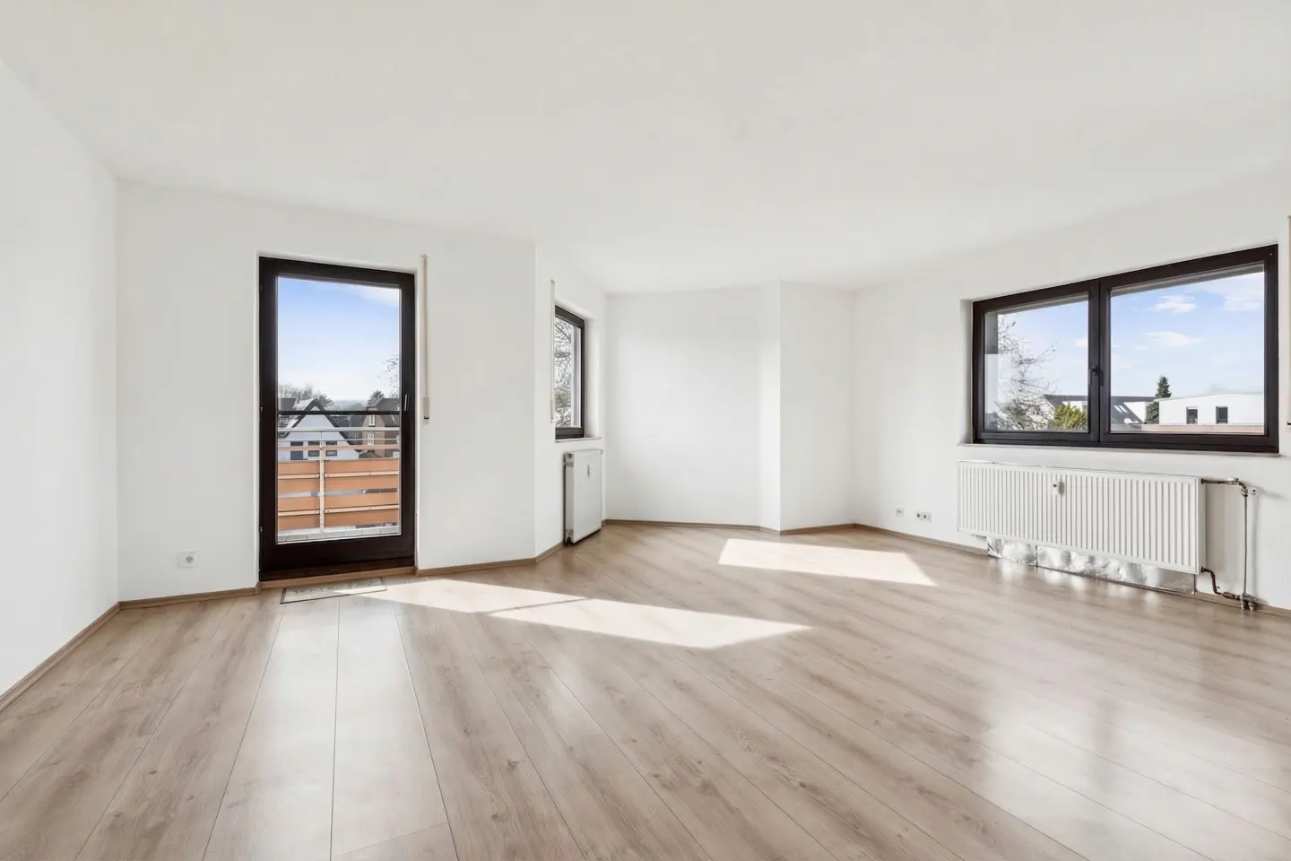 Bright, empty room with light wood floors, white walls, and black-framed windows. A door leads to a balcony. Radiators are under the windows.