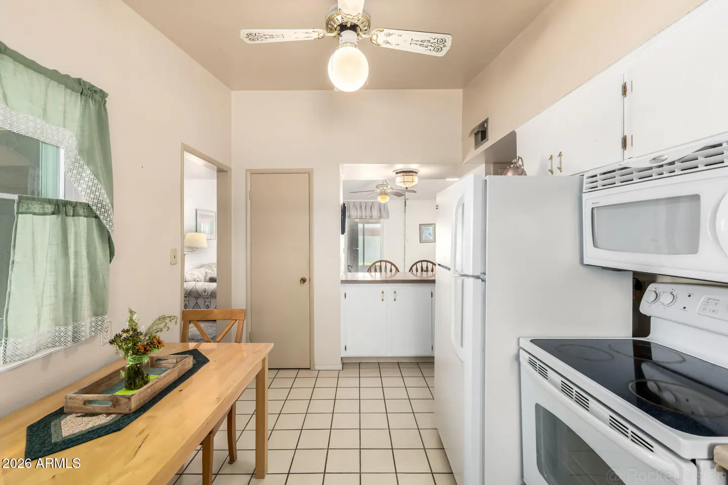 A bright kitchen with white appliances, tile floors, and a wooden table with a floral centerpiece. A doorway leads to another room.