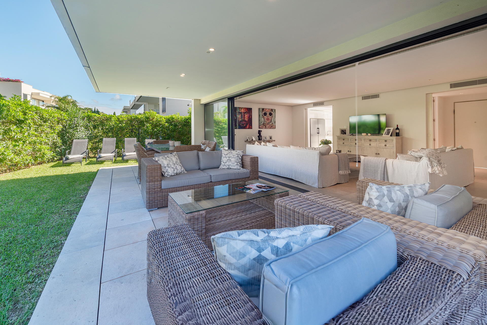 Modern patio with wicker furniture and glass coffee table, opening into a bright, contemporary living room with white sofas. [AI-generated text]