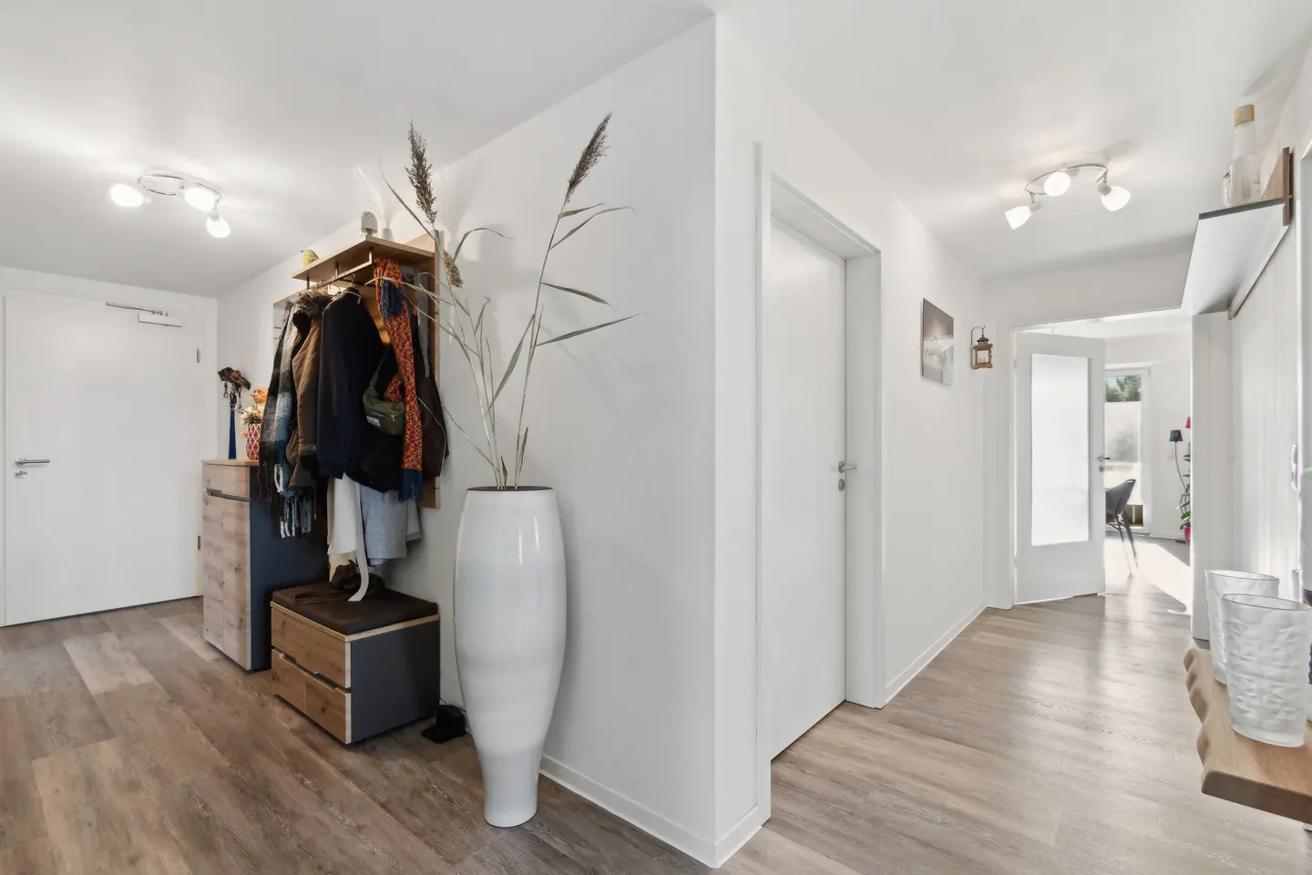 Hallway with wood-look floors, white walls, and doors. A coat rack with jackets, a cabinet, and a tall white vase with reeds are visible.