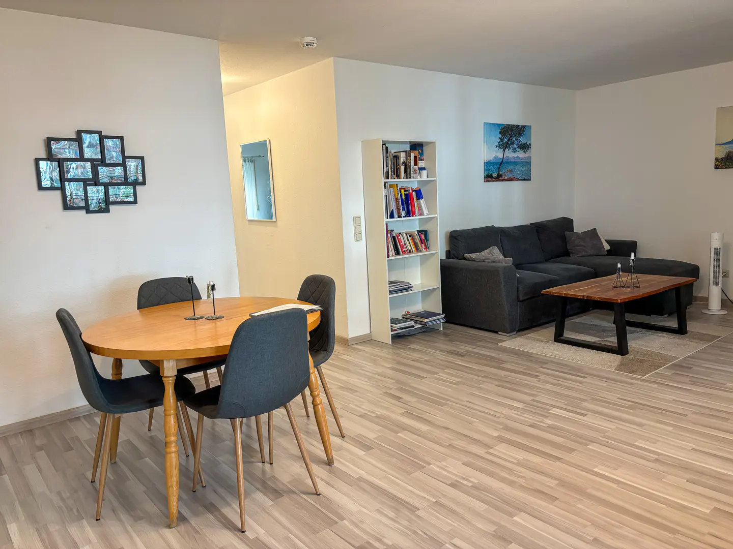 Open-plan living area with a wooden dining table, grey chairs, a bookshelf, and a dark grey sectional sofa on wood floors.
