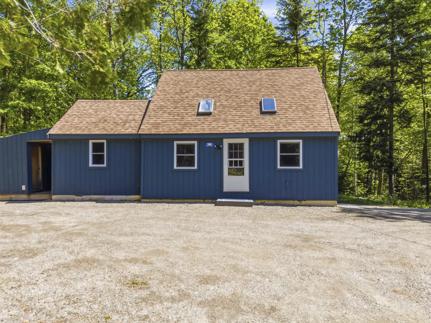 Blue cottage with a brown shingled roof, white trim, and two skylights, set against a backdrop of lush green trees.