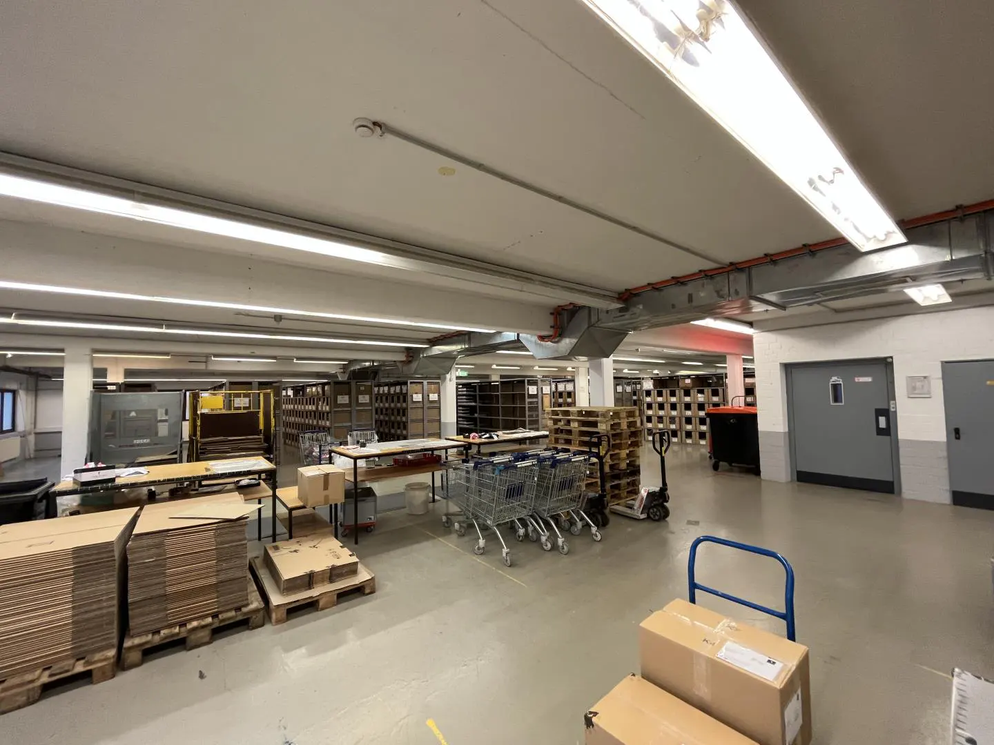 A large, brightly lit warehouse space with shelving, carts, and stacked cardboard boxes.