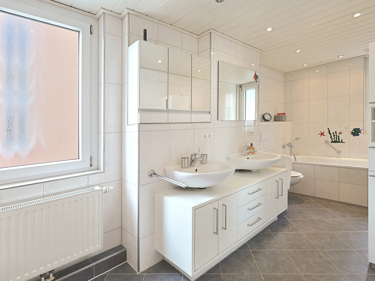 Bright bathroom with white cabinets, double sinks, and a large window. Gray tile floor and white tiled walls. Bathtub with fish decals.