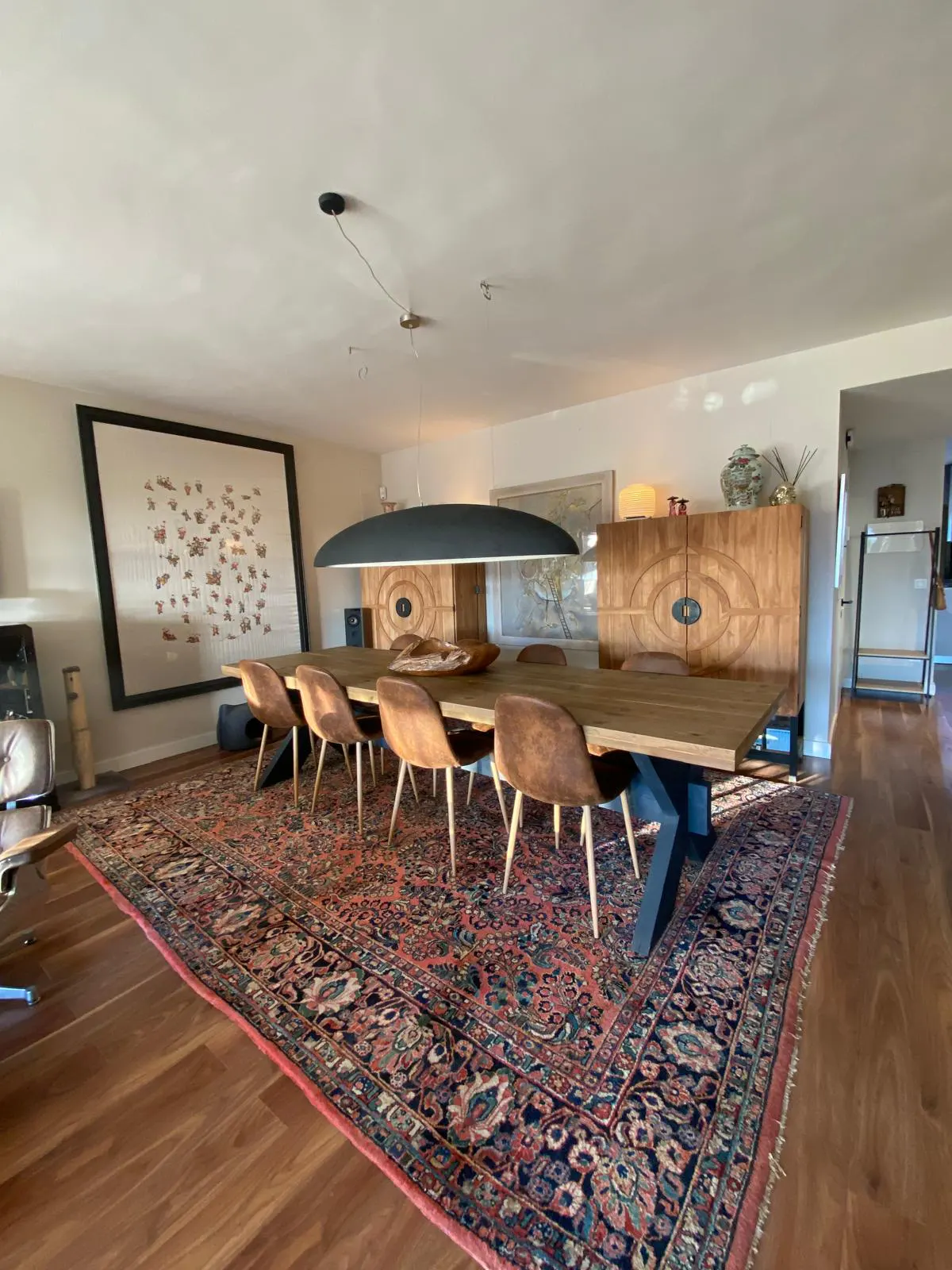 A dining room with a long wooden table, brown chairs, and a large patterned rug. A black pendant light hangs above the table.