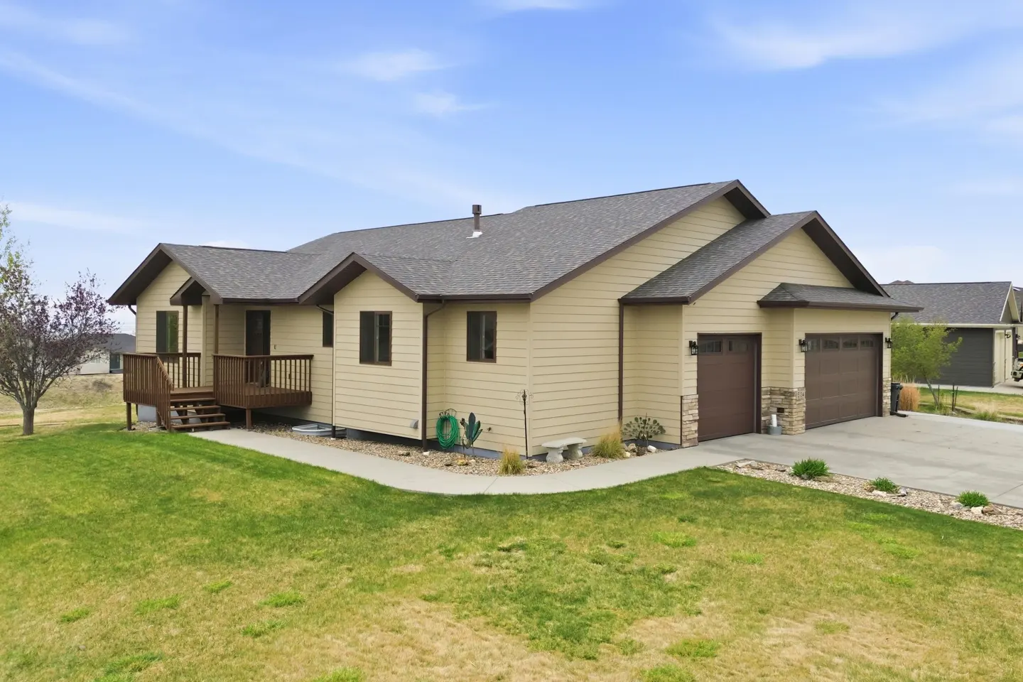 Tan single-story house with a brown roof, a wooden porch, and a two-car garage on a green lawn under a blue sky.