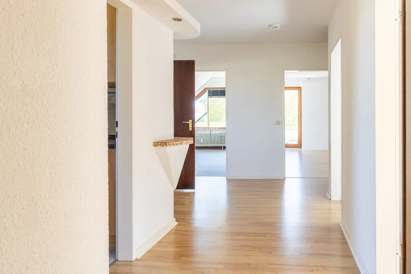 Hallway with light wood floors, white walls, and brown doors. A small shelf is mounted on the wall. Natural light streams in from windows in the distance.