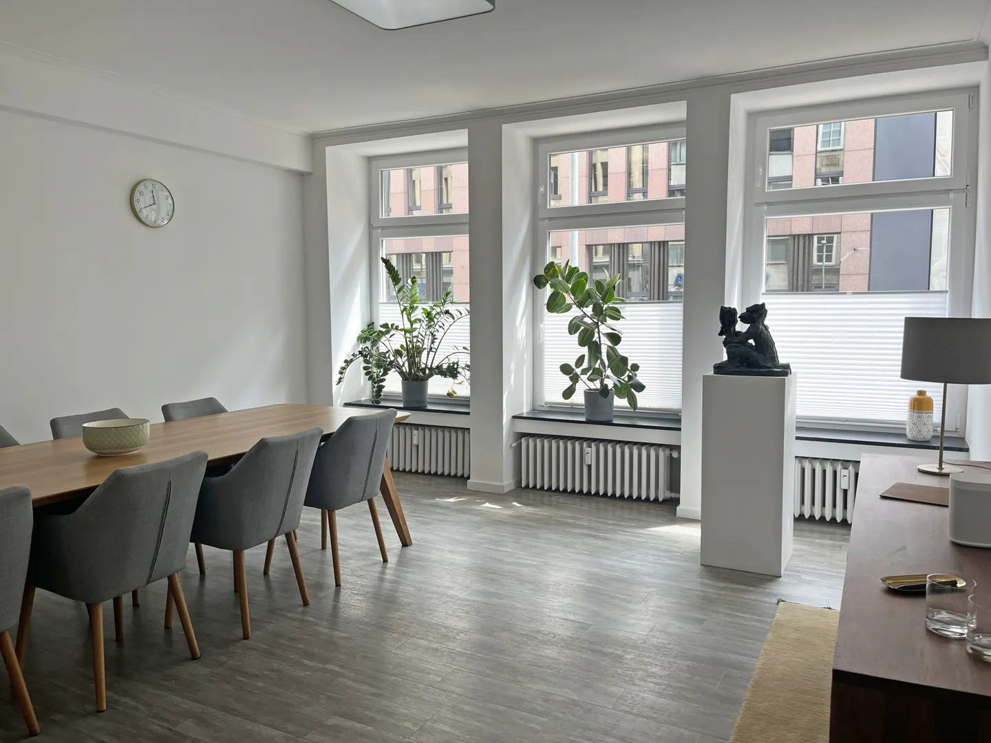 Bright dining room with a wooden table, gray chairs, and large windows with plants. A sculpture sits on a white pedestal.