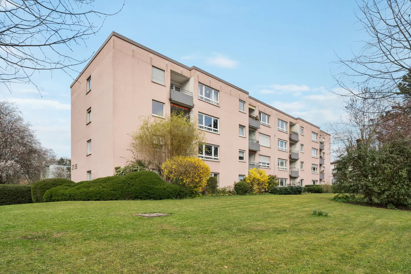 A pink apartment building with balconies, surrounded by green grass and bushes.