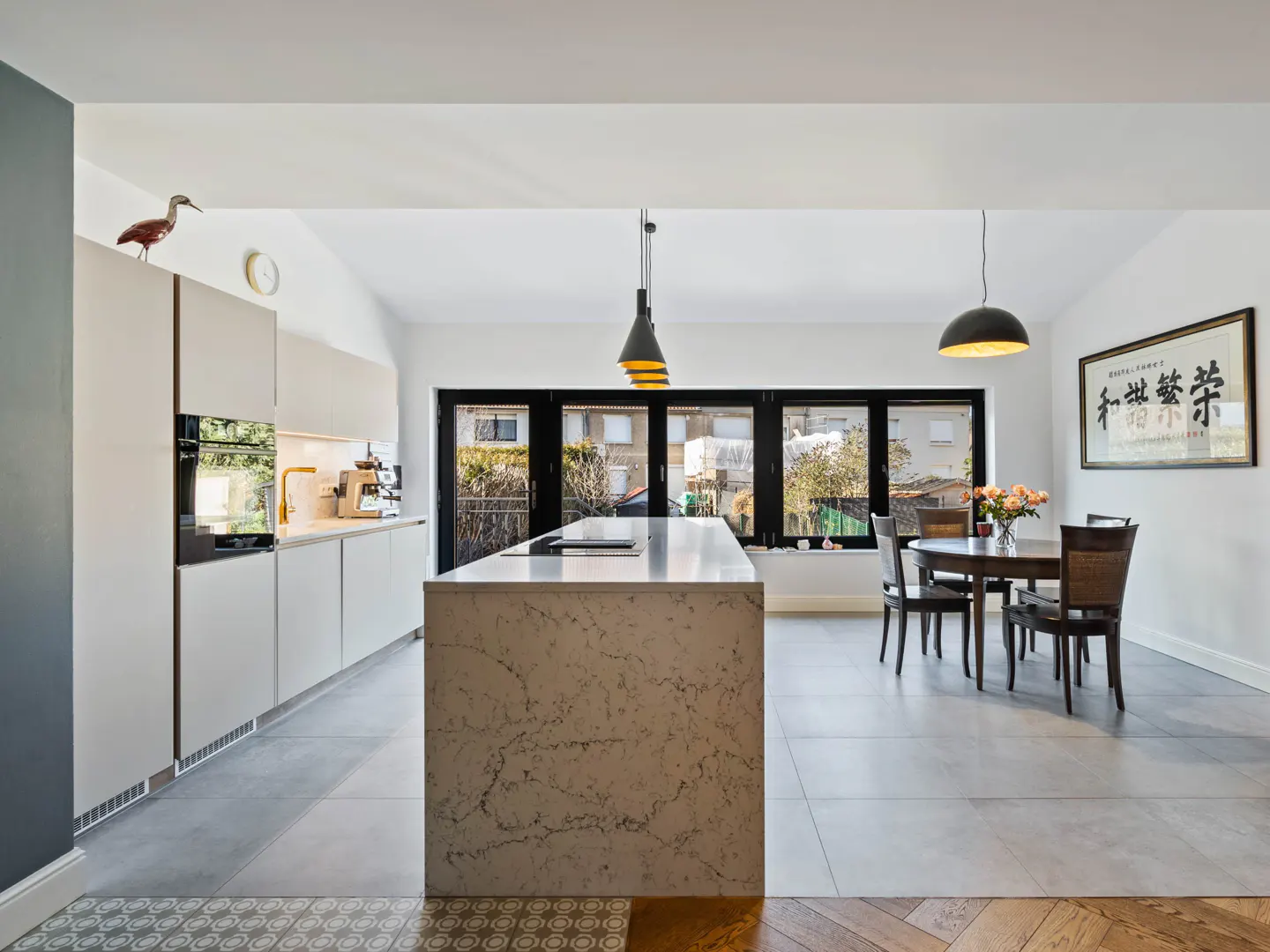Bright, modern kitchen with white cabinets, a marble island, and black-framed windows. A dining table sits near a framed print.