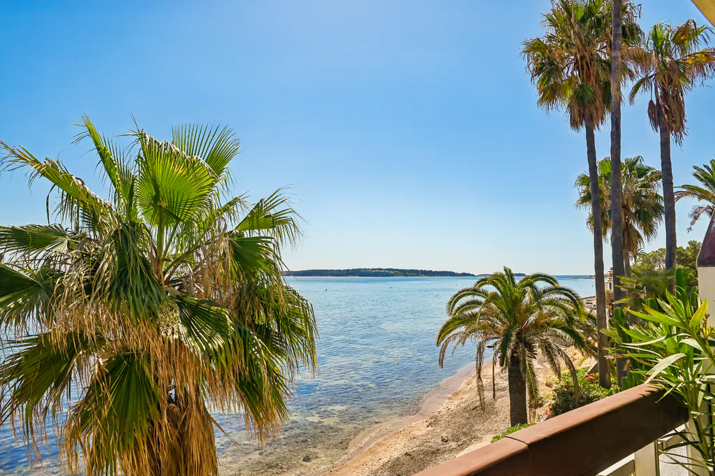 Balcony view of a sandy beach and turquoise sea under a clear blue sky, framed by palm trees.