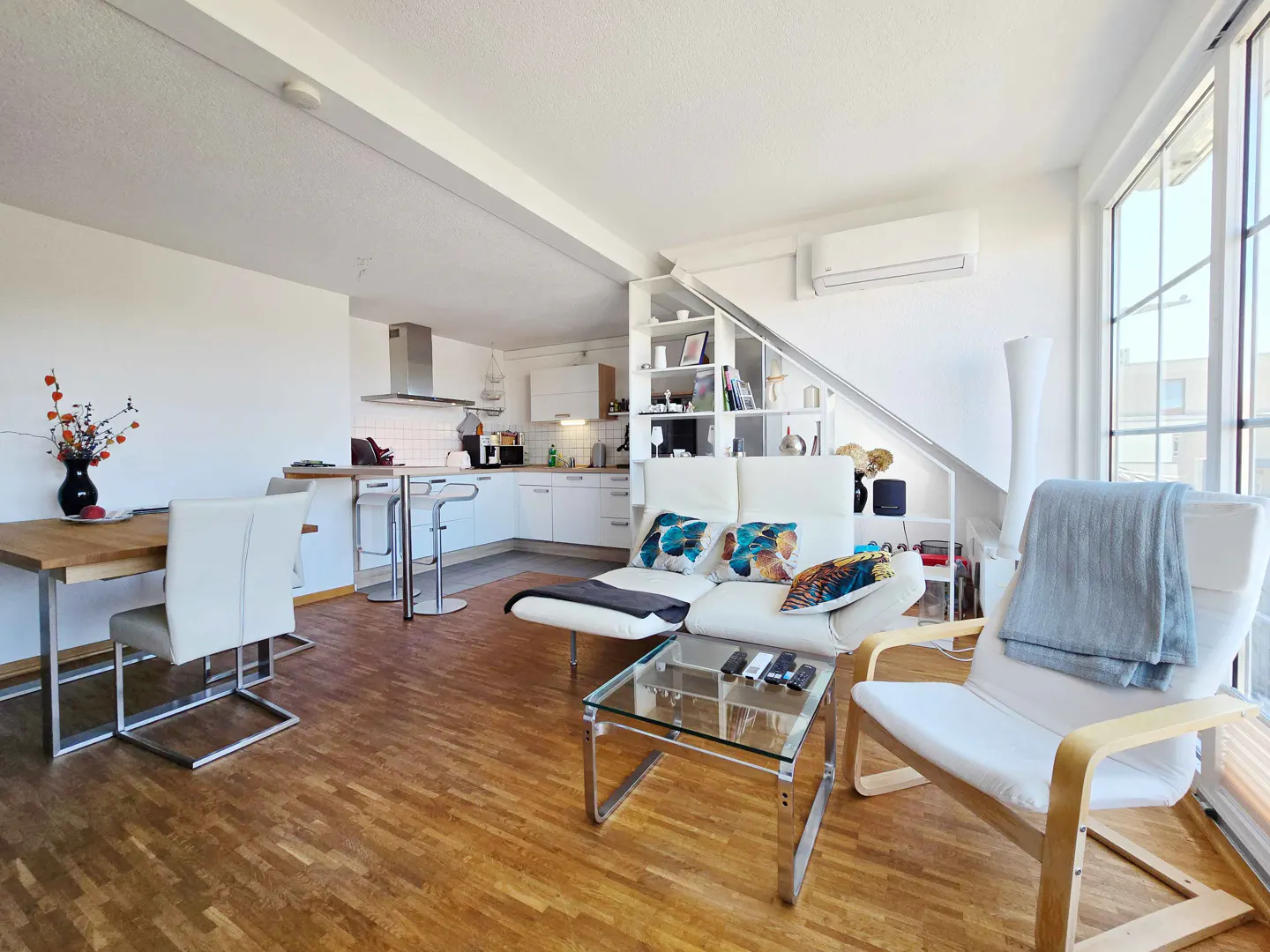 Bright, open-concept living space with wood floors, white walls, and large windows. A white sofa, chair, and glass table furnish the living area. A kitchen is visible in the background.