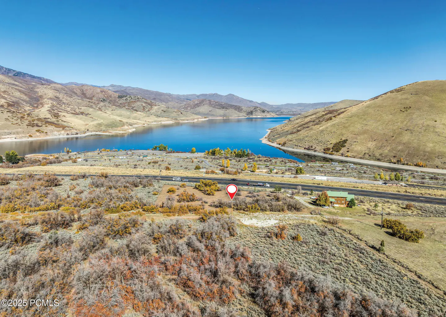 Aerial view of land with a red pin, a lake, mountains, and a road under a blue sky.
