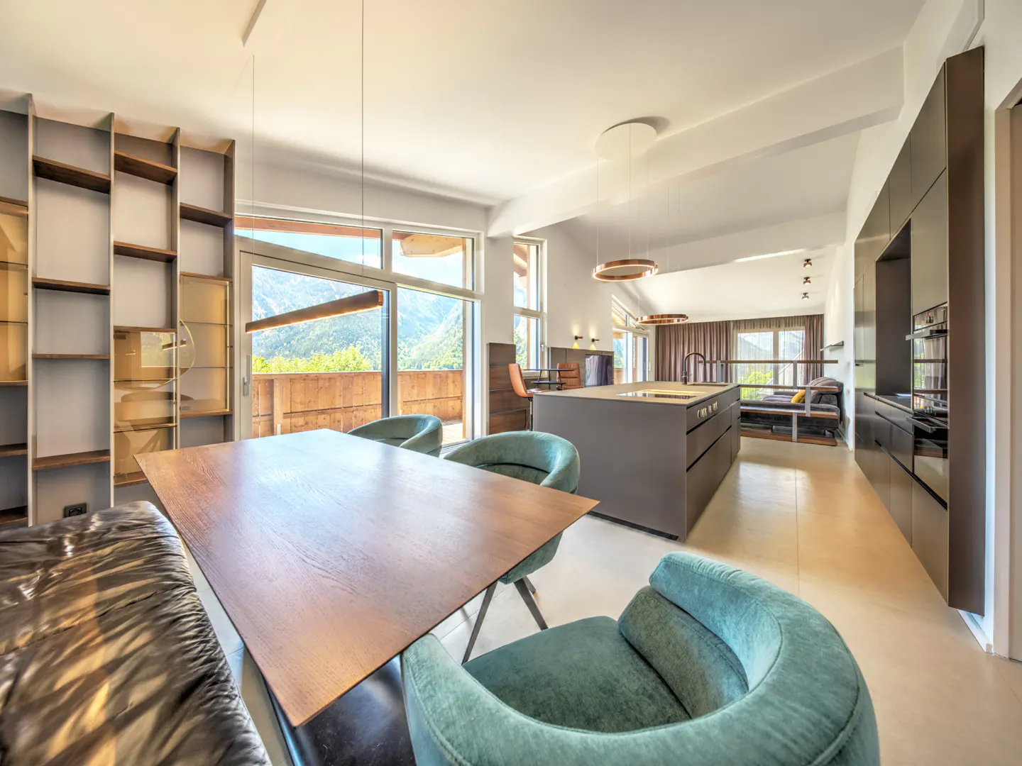 Modern open-plan kitchen and dining area with a wooden table, teal velvet chairs, and large windows overlooking mountains.
