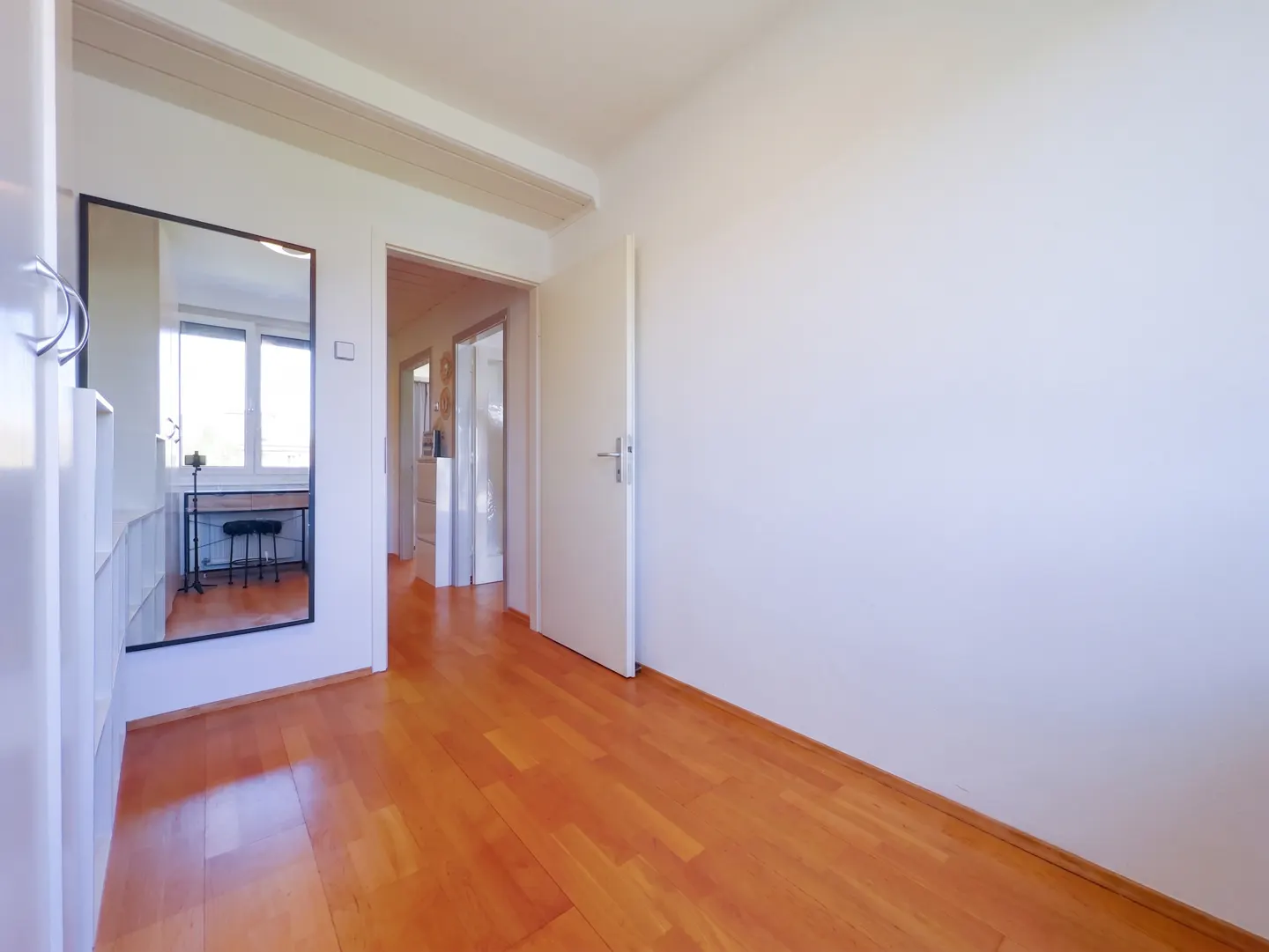 Bright hallway with wood floors, white walls, and a mirror reflecting a desk and window. A doorway leads to another room.