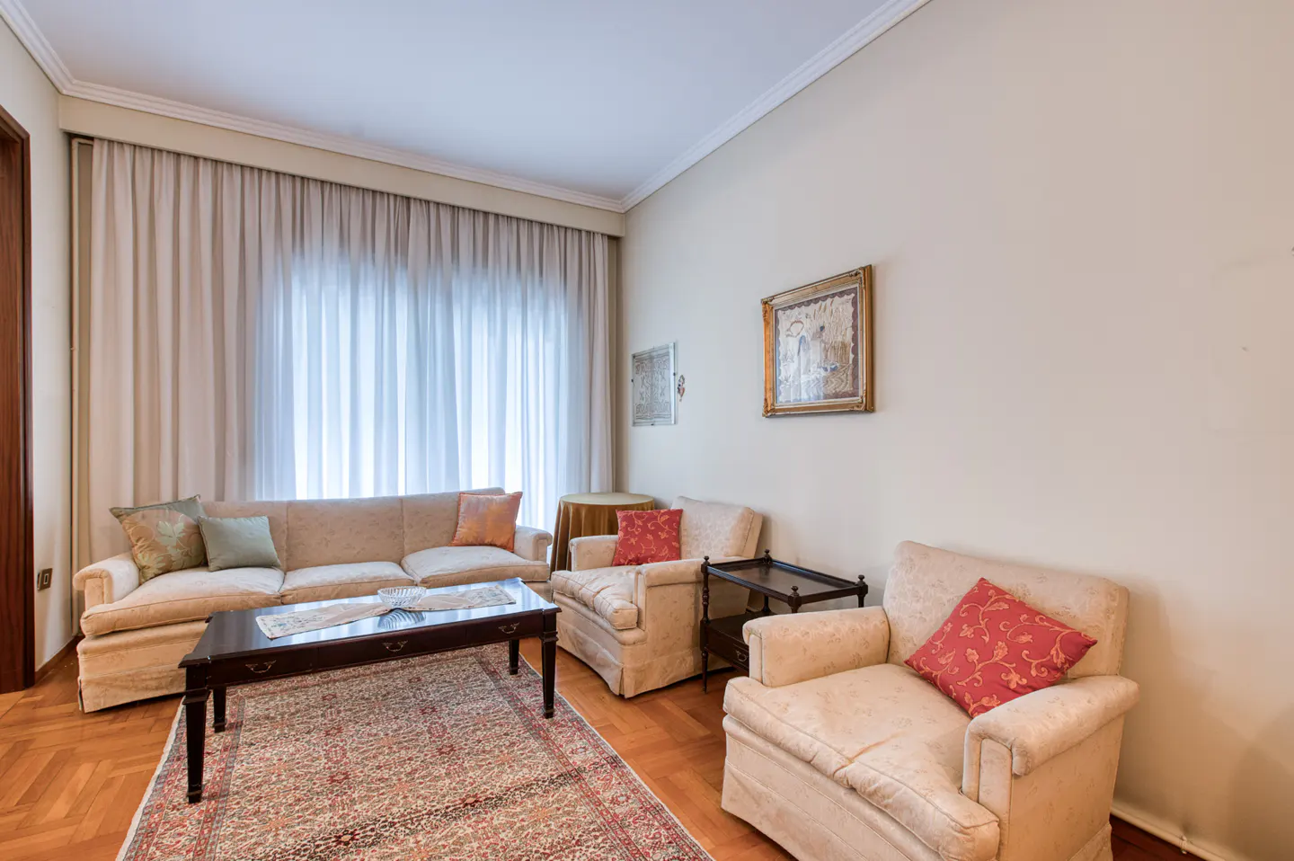 Living room with beige sofa, two armchairs, and dark wood coffee table on a patterned rug. Light walls, curtains, and framed art.
