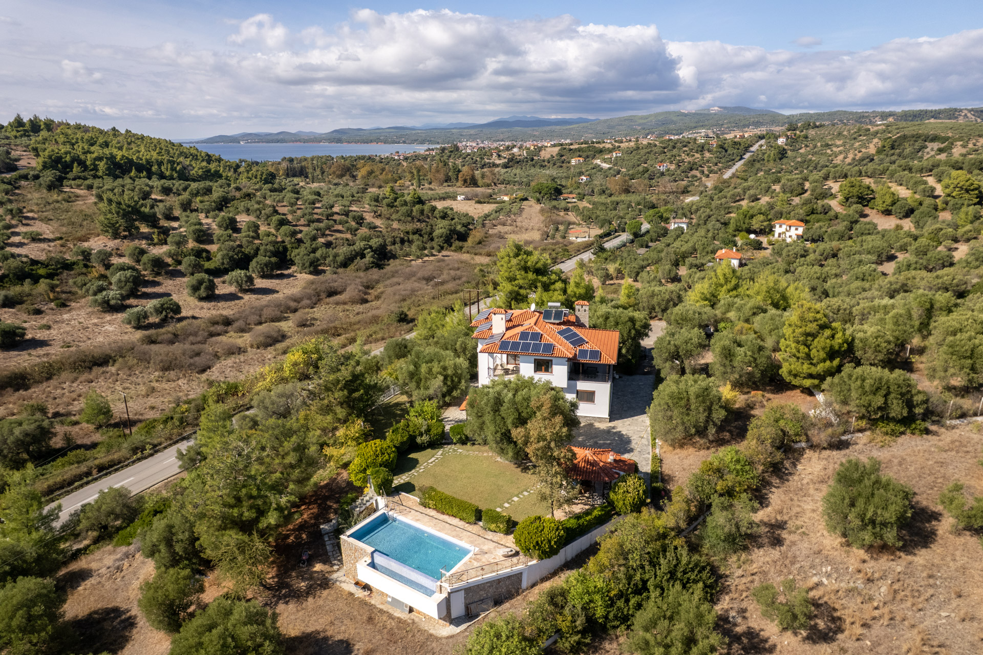Aerial view of a white villa with a red roof and solar panels, featuring a swimming pool, set in a lush, hilly landscape. [AI-generated text]