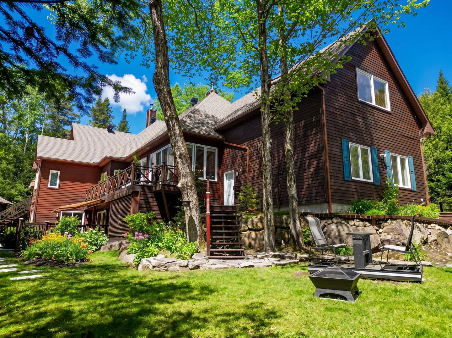 Exterior view of a brown, two-story house with a green lawn, trees, and blue sky. A fire pit and chairs sit on a stone patio.