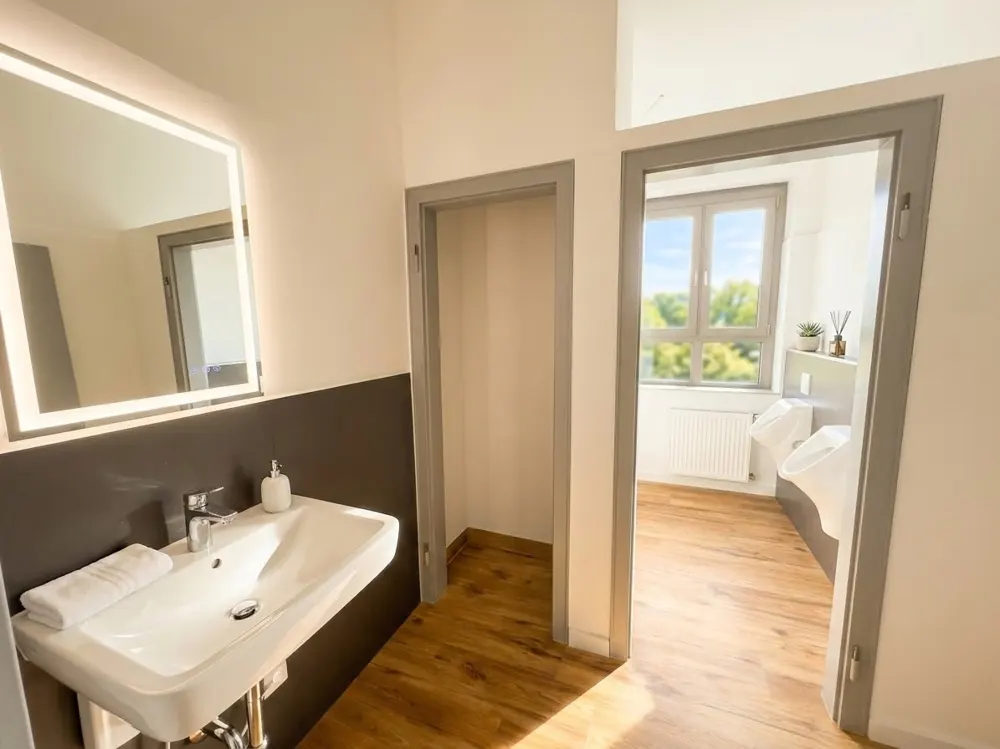 Modern bathroom with a white sink, illuminated mirror, and a doorway leading to a room with two urinals and wood flooring.