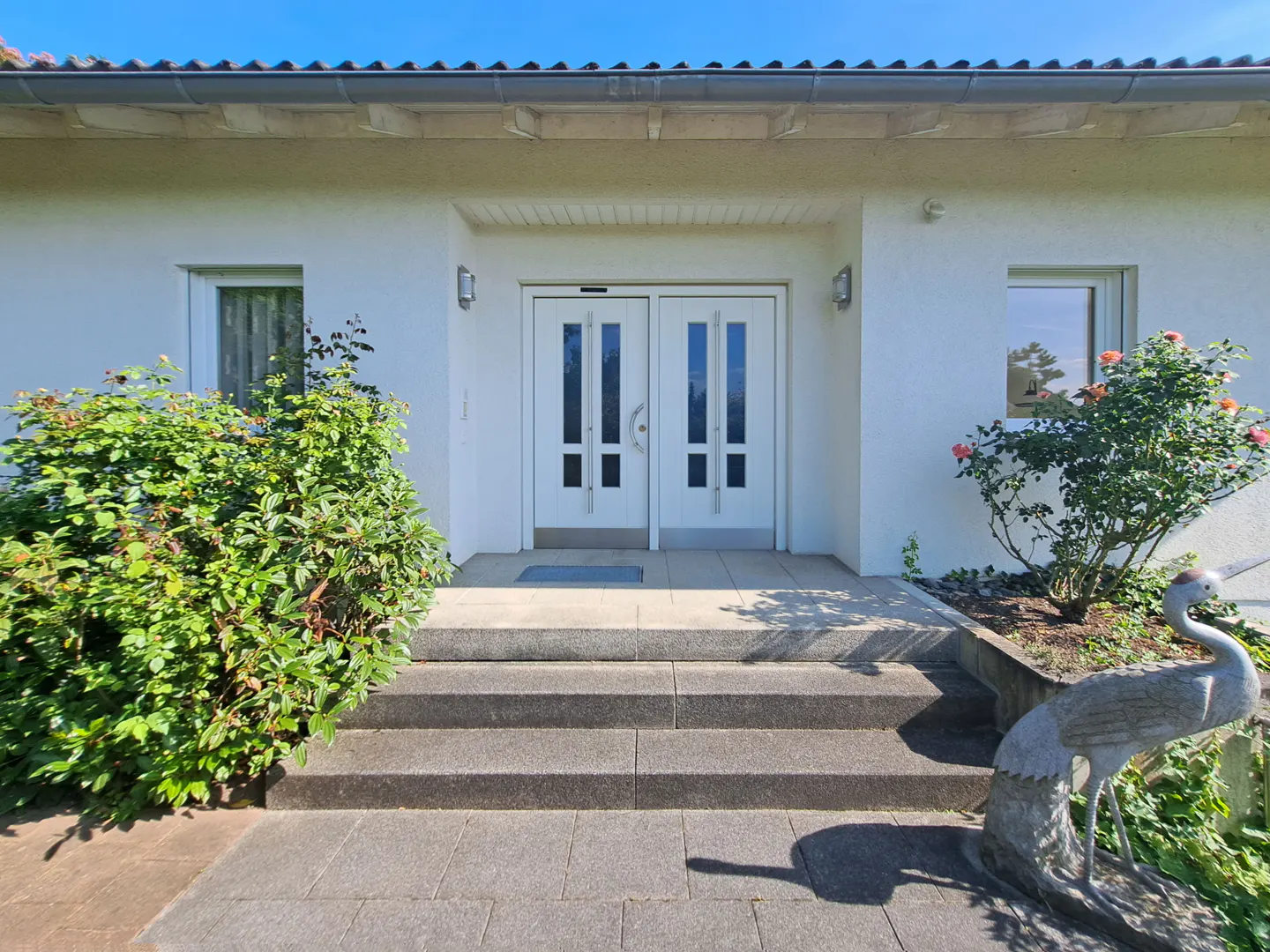 Front view of a white house with double doors, steps, and landscaping. A stone crane statue sits near a rose bush.