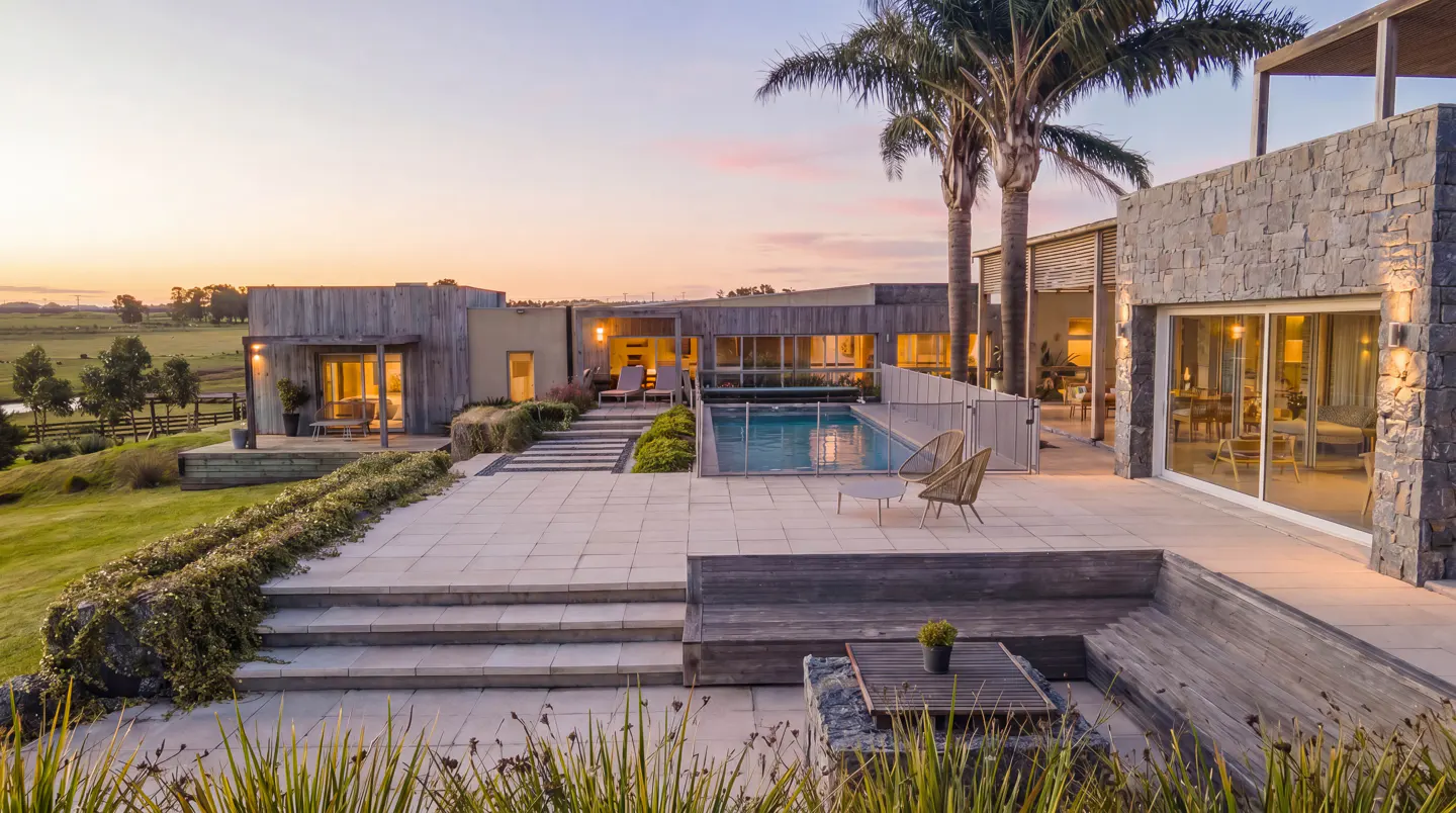 Modern home exterior with a pool, stone patio, and palm trees at sunset.