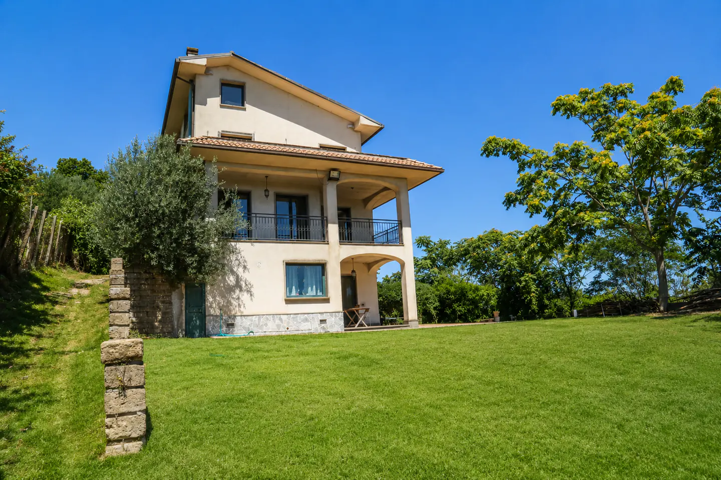 Two-story beige house with a balcony, green lawn, trees, and blue sky.
