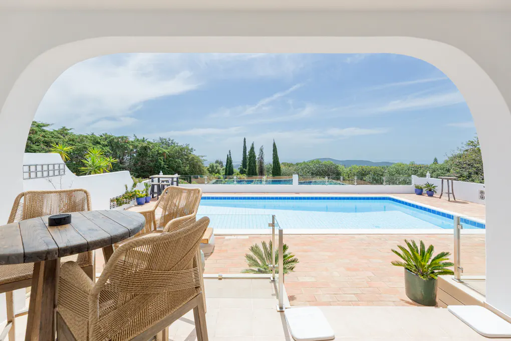 Outdoor patio with a round wooden table and four woven chairs overlooking a blue swimming pool and green landscape.