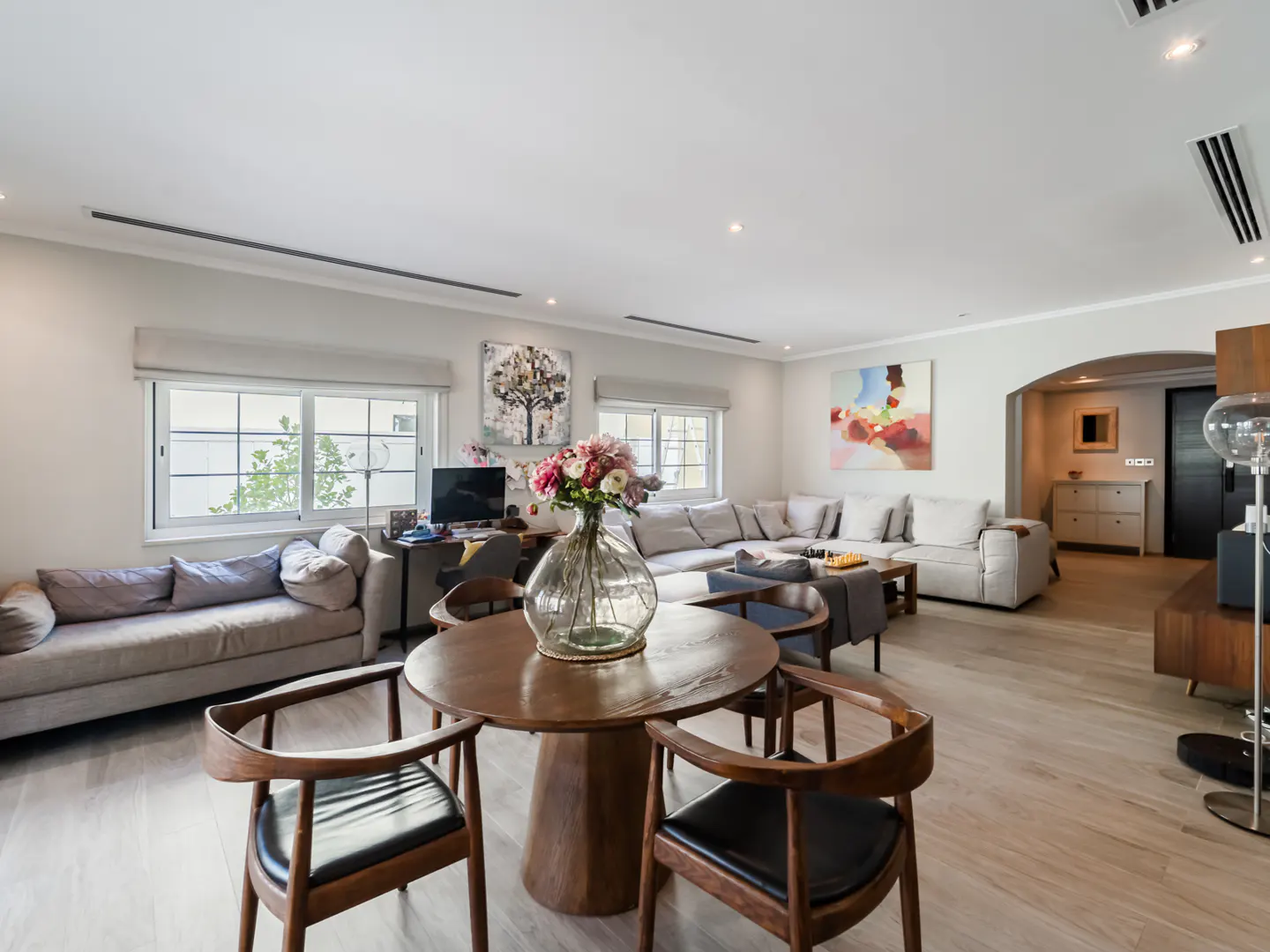 Bright living room with wood floors, a round table with flowers, and a large sectional sofa. Artwork hangs on the walls.