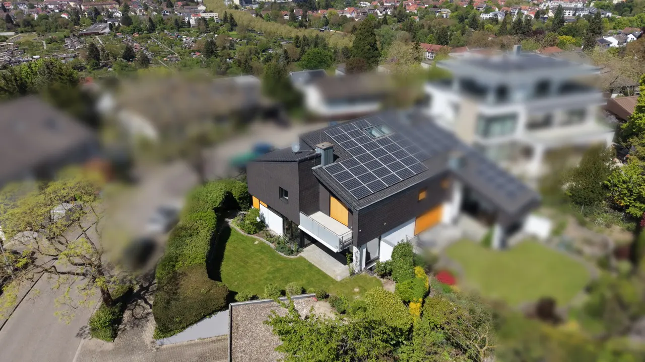 Aerial view of a modern dark-paneled house with solar panels on the roof, a small lawn, and a surrounding suburban area.